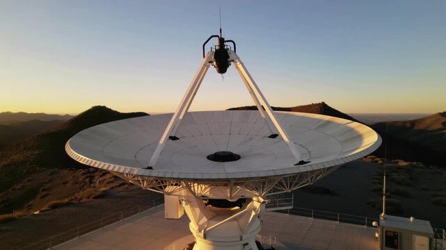 Large radio telescope dish antenna on a mountaintop at sunset with a clear blue and orange sky.