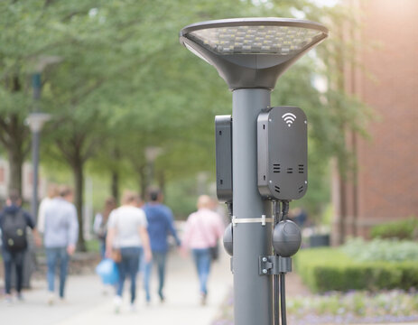 A focused medium shot of a smart streetlight on a campus walkway, equipped with WiFi transmitters and motion sensors, softly blurring pedestrians and greenery in the background.