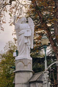 Lourdes, France - 19 November 2025: View of a pristine white angel statue with outstretched arm, juxtaposed against the muted autumnal hues of surrounding trees and a vintage lamp post.