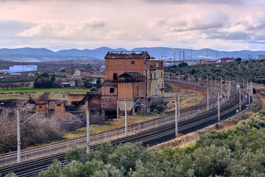 Puertollano, Spain - 17 December 2025: View of an aged brick structure, part of a former coal mine, standing beside curving train tracks, contrasting with the verdant foreground.