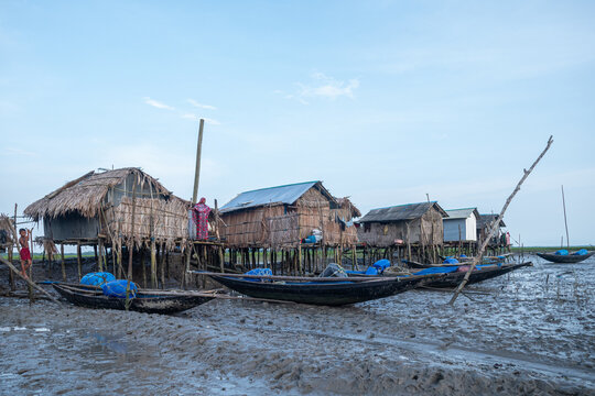 Khulna, Bangladesh - 12 July 2025: View of stilt houses with thatched roofs reflected in the muddy foreground, boats resting, under a pale sky.