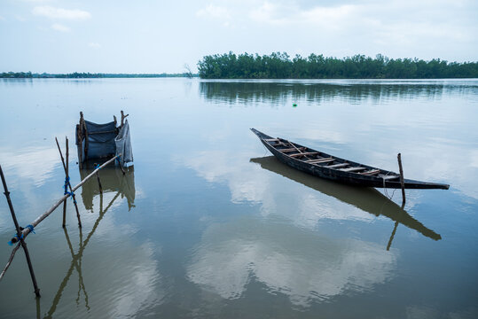 Khulna, Bangladesh - 12 July 2025: View of a traditional wooden boat and a fishing structure resting on calm waters reflecting the overcast sky.