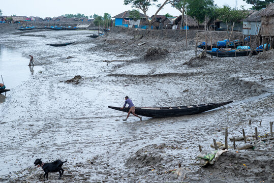 Khulna, Bangladesh - 13 July 2025: View of a lone figure struggling to move a boat through the thick, grey mudflats near the river, juxtaposed against the simple coastal village huts.