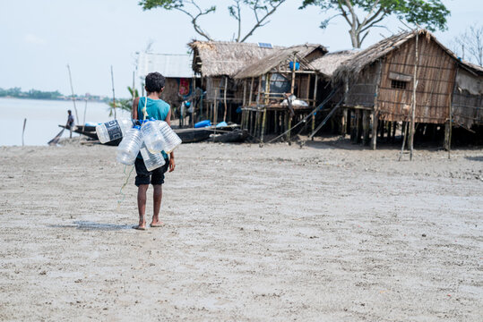 Khulna, Bangladesh - 13 July 2025: View of a young boy carrying plastic bottles along the muddy shoreline, leading towards stilted bamboo homes with thatched roofs under a pale sky.