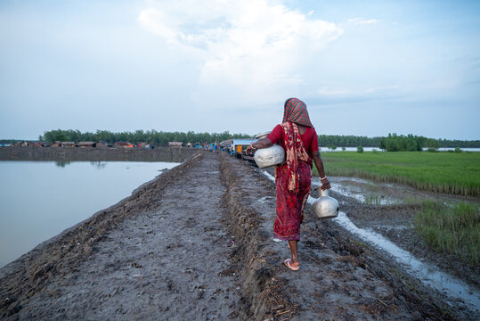 Khulna, Bangladesh - 12 July 2025: View of a woman in a red dress walking on a muddy path holding water containers, with flooded fields reflecting the sky.
