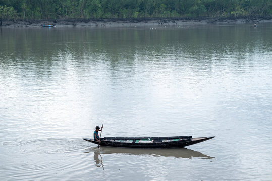 Khulna, Bangladesh - 13 July 2025: View of a lone figure rowing a narrow wooden boat across the calm, reflective river under the soft light of day, with dense greenery lining the distant shore.