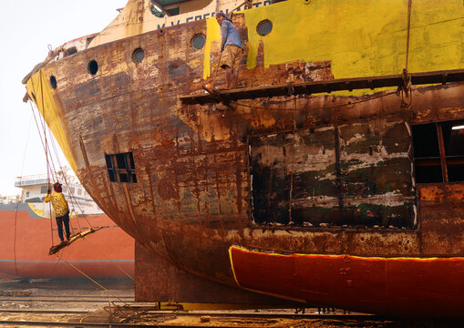 Keraniganj, Bangladesh - 11 February 2023: View of workers precariously perched on a rusted ship, amidst peeling paint and corroded metal, against the backdrop of the ship-breaking yard.