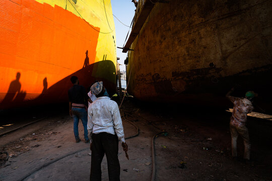 Keraniganj, Bangladesh - 28 November 2022: View of men dwarfed by towering, brightly colored ship hulls looming overhead, casting long shadows in the shipyard dust.