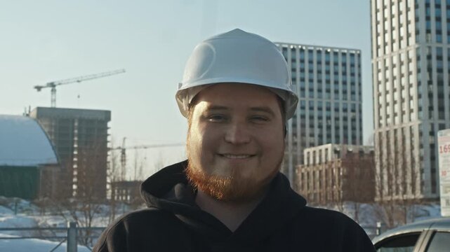 Portrait of builder against cranes and under-construction buildings in winter or early spring urban landscape.