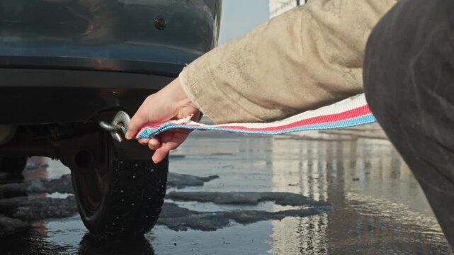 Driver checks reliability of tow cable hook attachment to vehicle's hitch ring on snowy parking lot by building.
