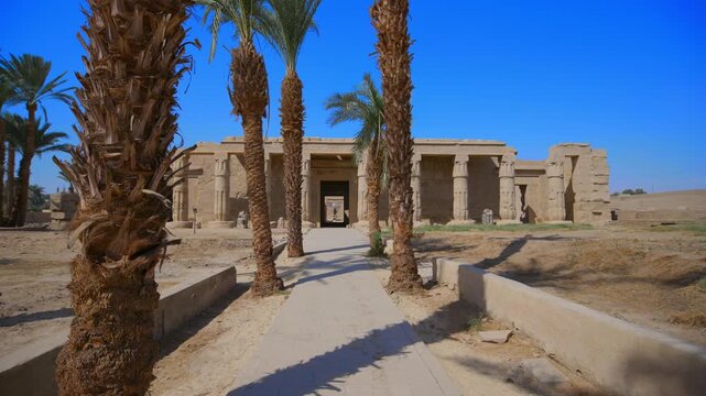 Seti I mortuary temple entrance with palm tree alley and ancient stone ruins in Luxor Egypt. Scenic pathway under vivid blue sky.