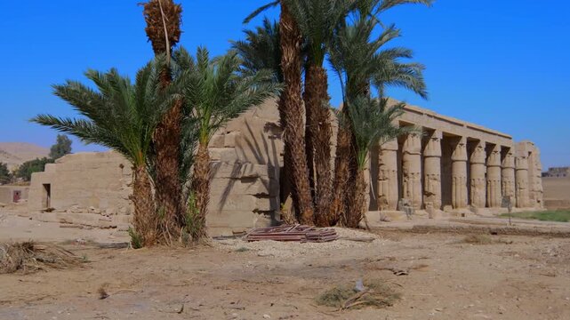 Seti I mortuary temple exterior with Osiride colonnade and palm trees in Luxor Egypt. Ancient sandstone ruins in desert landscape.