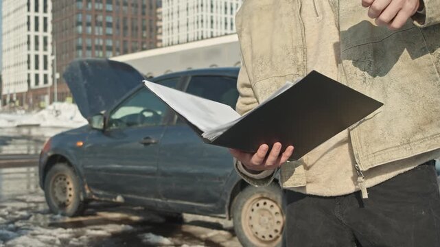 Winter car breakdown: driver dials insurance service holding documents and smartphone by black auto.