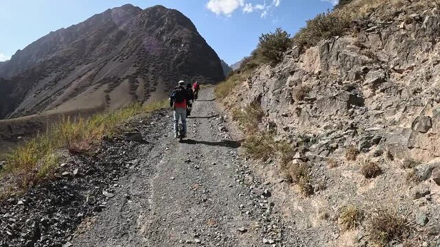 Three men riding electric unicycles along a gravel mountain road with rocks in Kyrgyzstan. The scene shows rugged terrain, natural surroundings, and off-road adventure.