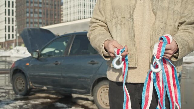 Driver in beige jacket holds colorful tow strap with two metal hooks by car with open hood.