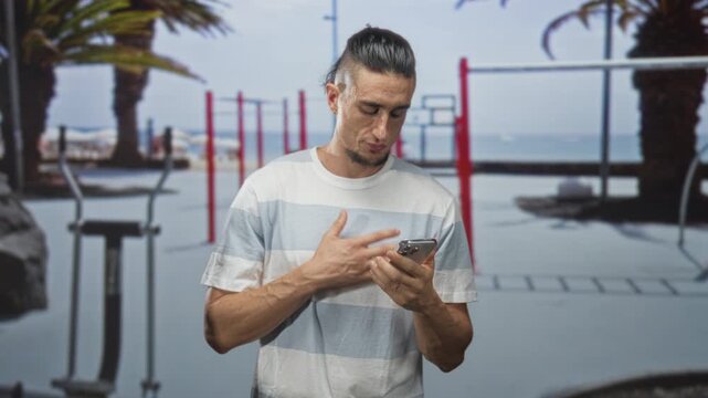 Man holding smartphone and tapping screen while looking down near outdoor fitness bars and palm trees on street; contemplation focus.
