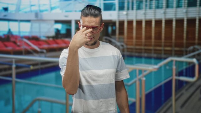 Man points finger to camera wearing red glasses and striped tshirt beside indoor pool railing in building; confidence boldness challenge.