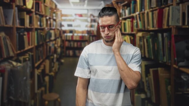 Man touching cheek with hand in a crowded bookstore aisle inside building, wearing red glasses and striped tee, eyes closed in discomfort; toothache pain concern.