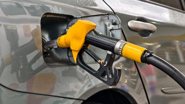 A yellow fuel nozzle is inserted into the gas tank of a gray car at a fueling station, highlighting the routine process of refueling a modern internal combustion engine vehicle for transportation.