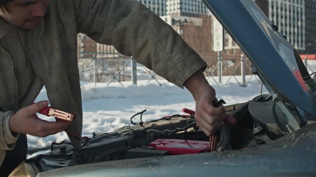Man charges battery old-fashioned way, attaching red and black jumper cables to car terminals for jump-starting dead winter battery.