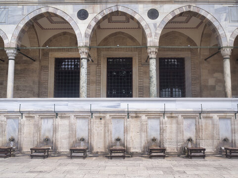 A symmetrical, wide shot of the elaborate Ottoman ablutions fountain (şadırvan) and stone facade with triple marble arches and columns at a historic mosque complex. Islamic architecture and culture.