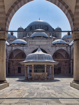 A symmetrical, wide-angle view of an ornate ablutions fountain (şadırvan) and the grand domes of an Ottoman mosque, perfectly framed by a large, central marble archway in the sunlit courtyard. Histor