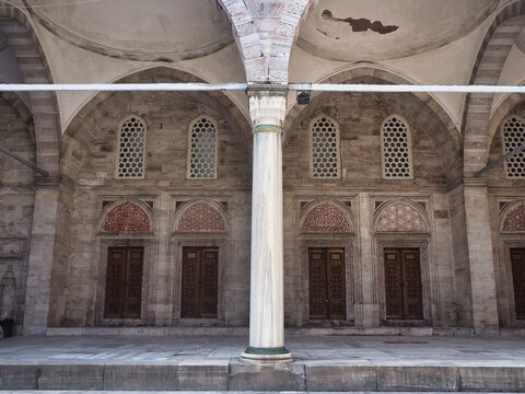 A striking symmetrical view of a mosque's inner courtyard gallery with wooden doors, decorative tympanums, arched windows with honeycomb grilles, and a central white marble column. Islamic ar