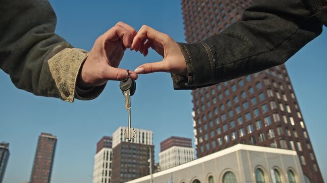 Couple makes heart shape with hands holding keys from purchased apartment in new multi-apartment building.