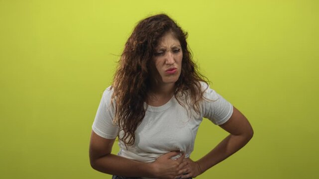 Young hispanic woman in white tshirt holding her stomach with hand on abdomen, grimacing and leaning forward in lime green studio; discomfort nausea.