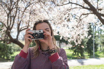 Fototapeta premium Young woman with a retro camera taking photos of a tree in bloom on a sunny spring day