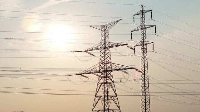 Aerial drone orbit around high voltage electricity pylons silhouette against sunset sky