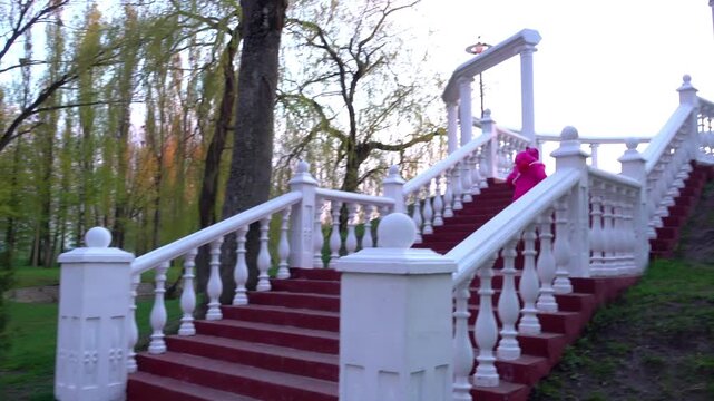 Beautiful girl in a pink jacket runs through the rotunda in the city park.