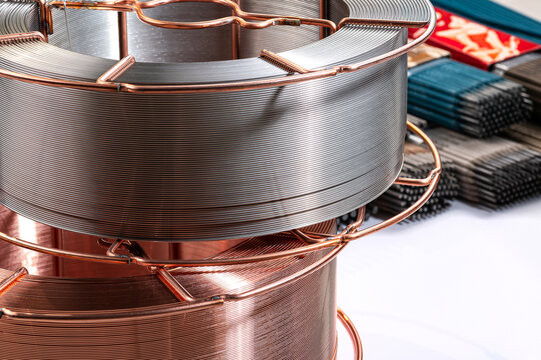 Steel welding wire spool on white background with electrodes. Large coil of silver steel welding wire on a metal reel with blurred electrodes.