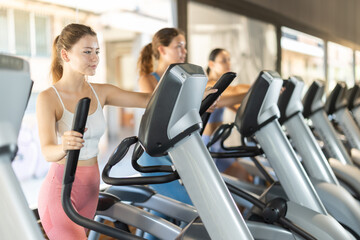 Fototapeta premium Smiling young girl training with elliptical machine in fitness hall during workout session