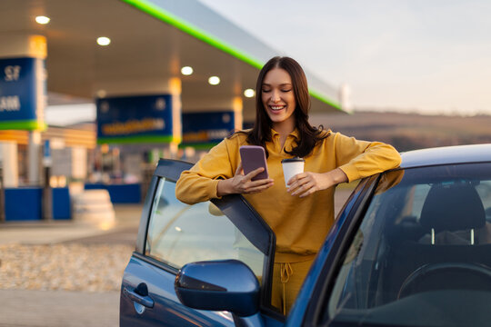 Lady using cellphone and drinking coffee while stopped at gas station, standing near her car, taking break and checking route on gps or texting