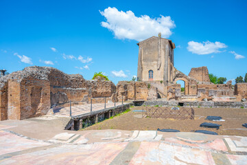 Fototapeta premium Visitors explore Casina Farnese at the Roman Forum in Rome, Italy. The historic site features ancient ruins and remains surrounded by blue skies and clouds. People walk around and take pictures.