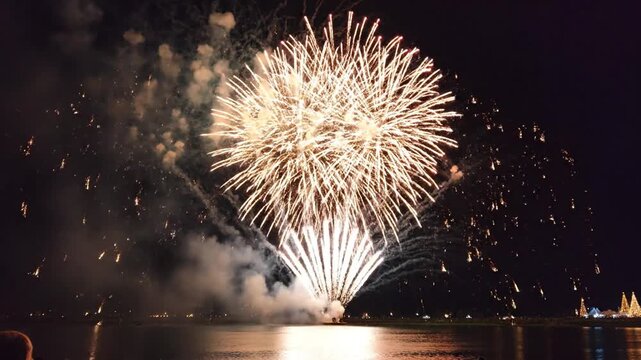 Fireworks exploding over water at night, reflecting colorful lights, with a city skyline in the background, possibly for celebrations or festivities