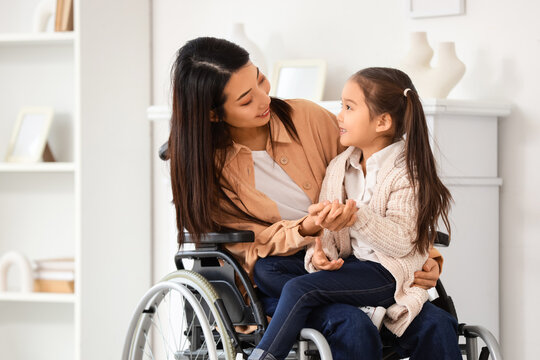 Young Asian mother in wheelchair with her little daughter holding hands at home