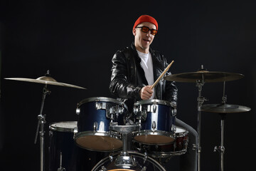 Obraz premium Smiling man in hat and sunglasses playing drums with drumsticks on black background