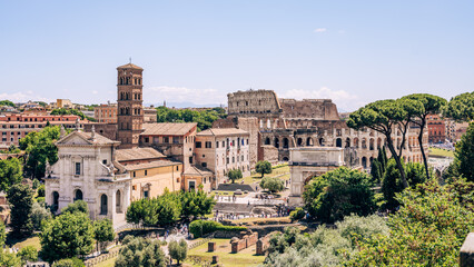 Fototapeta premium Visitors walk through the Roman Forum with views of the Colosseum in the background. People explore ancient sites under a clear blue sky during the day.