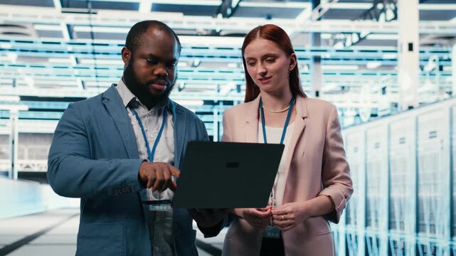 Engineering team in server hub working together with laptop to do maintenance tasks on machines. Teamworking colleagues in modern high tech data center configuring server settings, camera B