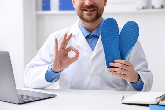 Smiling orthopedist in medical coat with foot insoles showing ok gesture at white desk in clinic, closeup