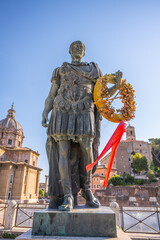 Obraz premium A statue of Julius Caesar stands tall at the Roman Forum in Rome. This historical site attracts many visitors. A wreath hangs on the statue as the sun shines brightly in the sky.