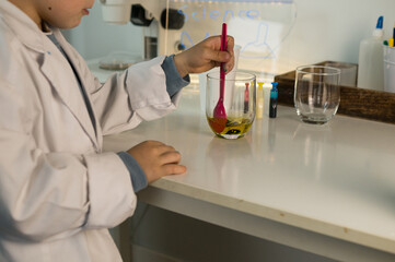 Fototapeta premium A young student uses a pink spatula to mix oils and colors in a glass, surrounded by art and science supplies. 