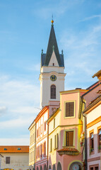 Fototapeta premium The Church of Our Lady the Queen and St. Jilj rises above colorful buildings in Trebon. The sky is clear with sunlight shining on the tower, marking an evening scene in the town.
