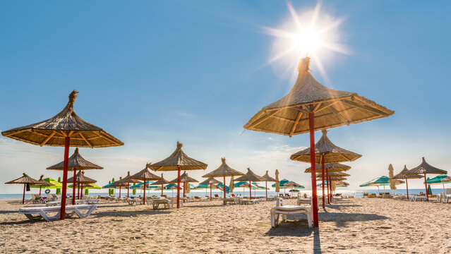 Sandy beach with rows of straw umbrellas on the Black Sea resort of Mamaia, Romania, in the morning after the sunrise, with the sun shining above the Black Sea.