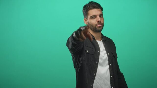 Man with beard, finger to temple while looking at camera, wearing white tshirt and black jacket on green studio backdrop; confidence intent focus.