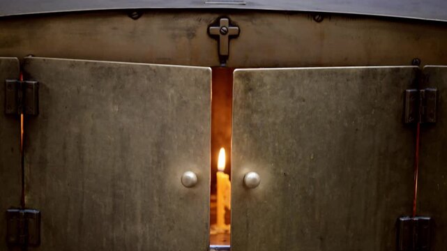 A candle burns softly, casting warm light through the gap of heavy metal doors