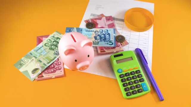 A hand drops a coin into a pink piggy bank surrounded by Ghanaian Cedi notes, a calculator, and a pen on a yellow surface, symbolizing personal savings, budgeting, and financial growth in Ghana.