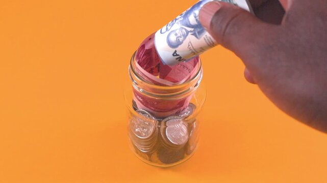 A hand places rolled 1 and 5 Ghana Cedi banknotes into a glass jar filled with coins. Set against an orange backdrop, it symbolizes saving money and financial growth using Ghanaian currency.